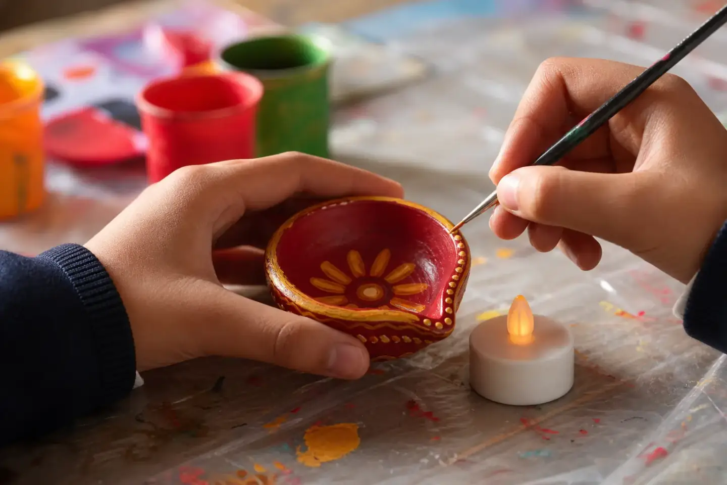 A pupil's hands steadying a painted terracotta diya while the end of a brush dots a pattern around the rim, little gems on the table beside it.